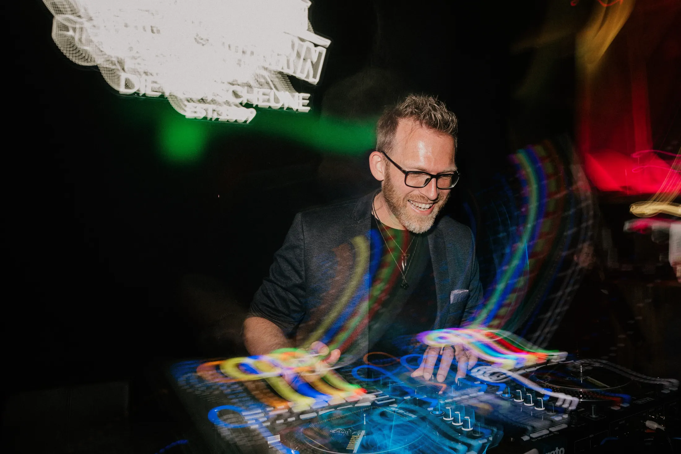 DJ Airbeck lacht am Pult, bunte Lichtspuren im Club